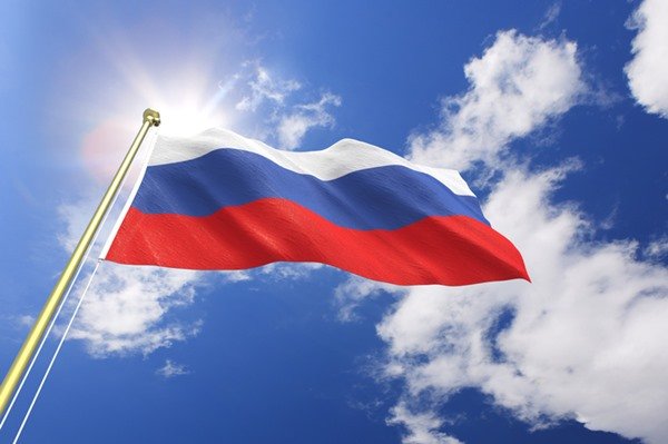 La imagen muestra la bandera de Rusia bajo el cielo con nubes y un sol brillante detrás - Metropolis