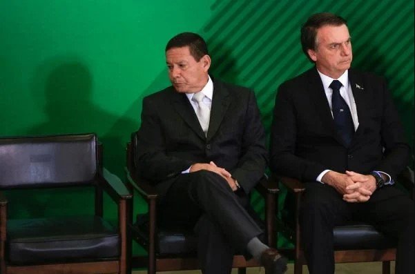 Bolsonaro e Mourão aparecem em evento no Planalto sentados.  Eles usam terno e gravatas para os lados opostos
