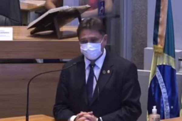 Homem de máscara, terno e gravata, falando em microfone