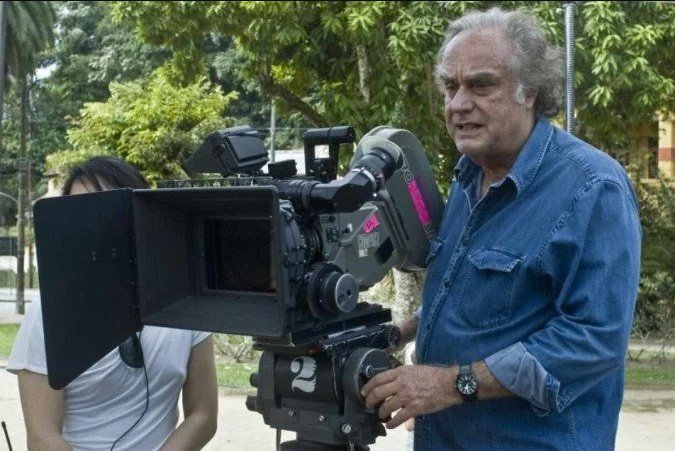 Na imagem colorida, um homem está posicionado à direita. Ele usa roupa jeans e está segurando uma grande camera