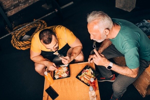 In the color image, two men occupy the center of the image.  They are eating from black plates and wear yellow and green blouses.