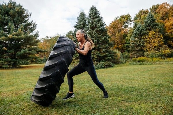 In the color image, a lady is positioned in the center.  She wears dark clothes and pushes a big tire.