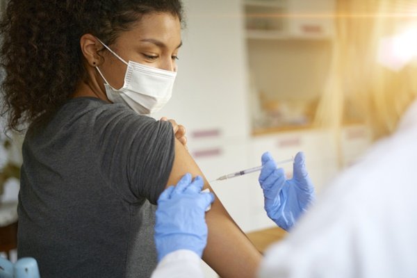 Na imagem colorida, uma mulher está posicionada no centro. Ela está com o braço de lado enquanto alguem aplica uma vacina