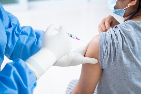 Na imagem colorida, uma mulher está posicionada no centro. Ela está com o braço de lado enquanto alguem aplica uma vacina
