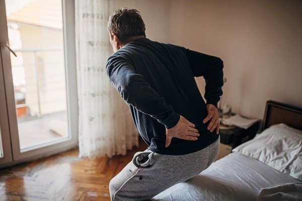 Pessoa com as duas mãos na parte inferior das costas. A pessoa está com os joelhos flexionados. Usa calça cinza e camiseta de manga longa na cor azul- Metrópoles