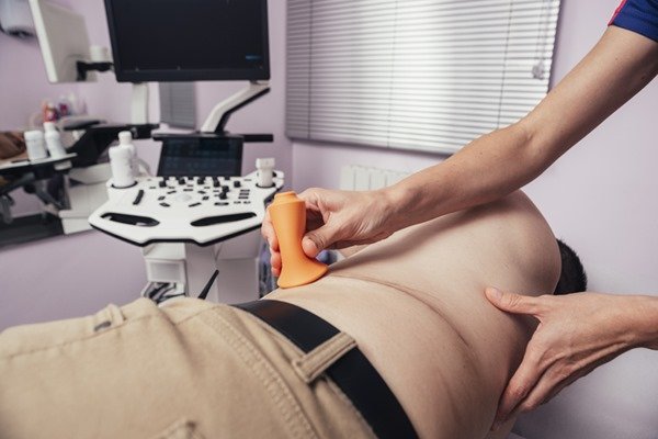 Pessoa fazendo exame na parte inferior da barriga em uma espécie de sala de clínica. A pessoa usa calça bege, está sem blusa e está deitada em uma mapa hospitalar - Metrópoles