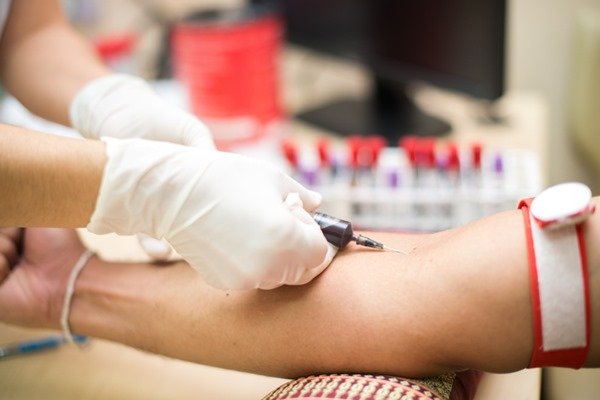 exame de sangue sendo realizado. Uma pessoa com pele clara segura uma seringa sobre um outro braço que também tem a pele clara