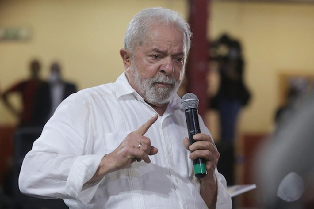 O ex-presidente Lula com um microfone na mão fala ao público. Na foto ele usa camisa social branca e ao fundo, desfocadas, aparecem outras pessoas - Metrópoles