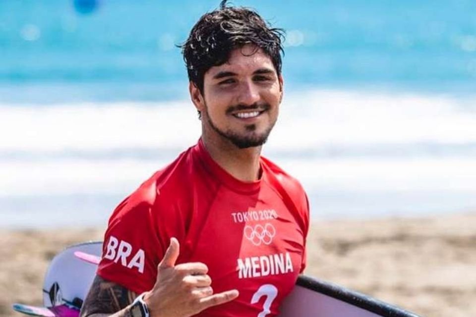 Na foto, Gabriel Medina aparece na beira da praia durante as Olimpíadas de Tóquio. O surfista segura dua pranca branca com traços azuis veste uma camisa vermelha. Ele sorri para a câmera e faz um sinal com a mão de hang loose