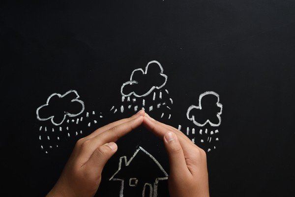 Mão protege casa de chuva. A casa e as nuvens com chuva estão desenhadas em quadro com giz-Metrópoles