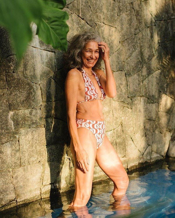 Mulher branca e idosa com biquini estampado branco e marrom. Ela está em uma piscina rasa.