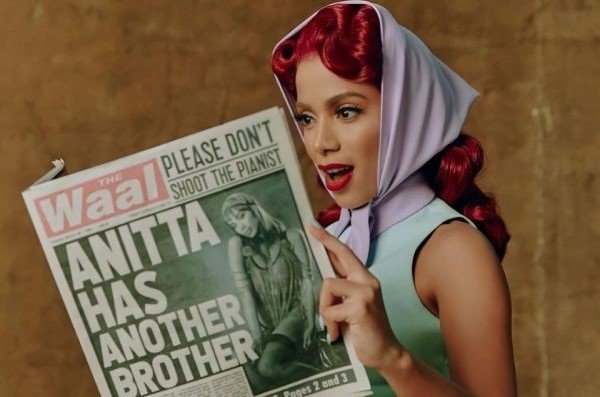 Singer Anitta posing for a photo.  She wears green clothes, a lilac scarf on her head and looks puzzled at a newspaper - Metropoles