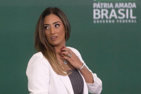 Na cerimônia de lançamento do projeto "Brasil para Todos", a ministra Flávia Arruda olha para o lado, com a mão no pescoço. Ela usa uma roupa branca e preta - Metrópoles