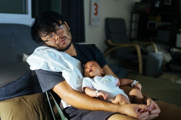 Homem sentado em cadeira cochila enquanto segura um bebê no colo-Metrópoles