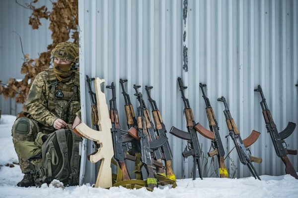 Soldado ucraniano participa de treinamento militar de civis. Ele usa uniforme militar, o chão está coberto de neve e o soldado se agaixa em muro de metal com armas AK-47 escoradas - Metrópoles