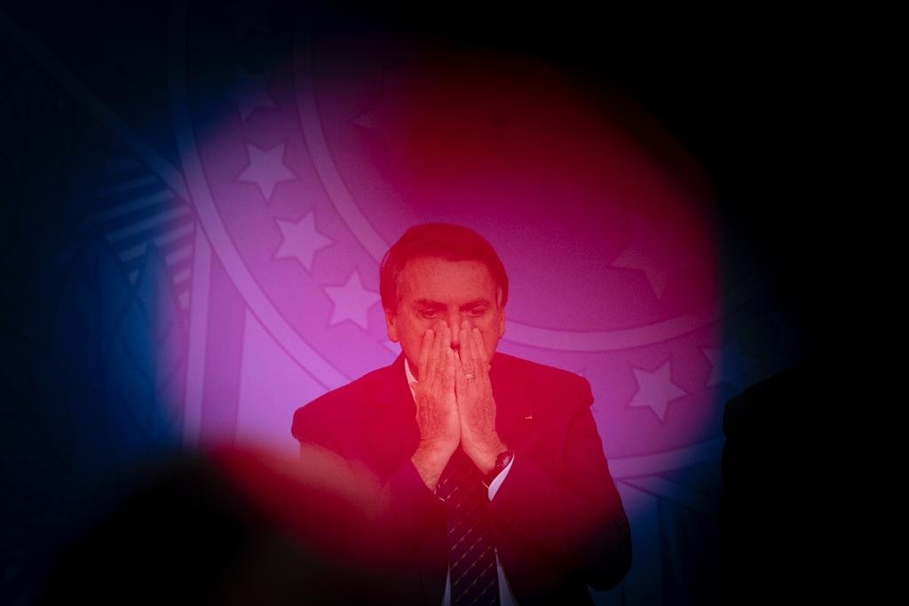 Bolsonaro sob luz vermelha na cerimônia de lançamento da Carteira de Identidade Nacional no Planalto. Ele está com as mãos no rosto - Metrópoles