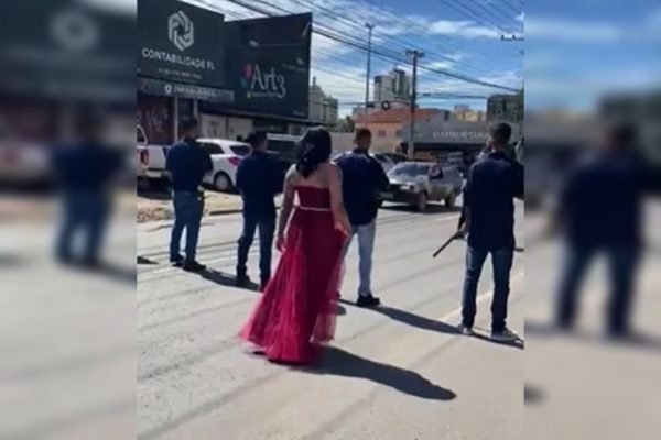 Mulher de vestido vermelho, de costas, e homens armados em pista