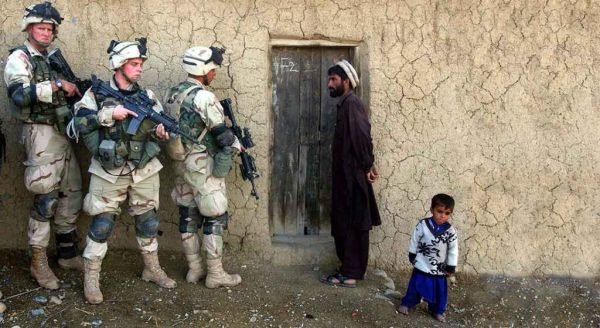 Militares da Otan fortemente armados ao lado de pai e filho afegãos em frente a parede de barro, na guerra do Afeganistão - Metrópoles