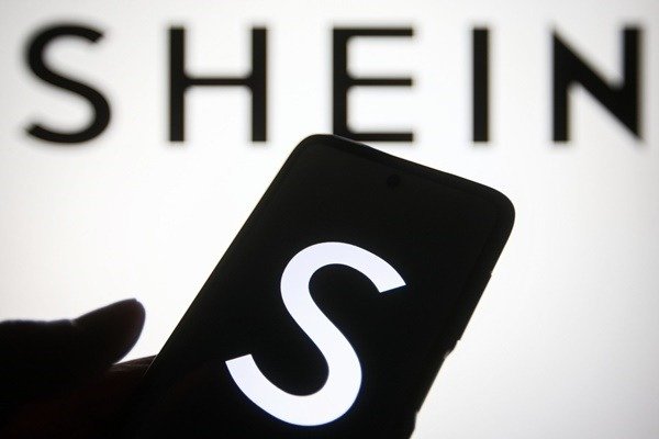 Na imagem, o logotipo da Shein está ao fundo enquanto uma mão segura um celular com S na tela 