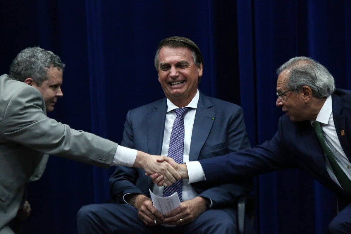 Bolsonaro ri sentado entre o ministro da economia Paulo Guedes e outro homem, que dão as mãos - Metrópoles