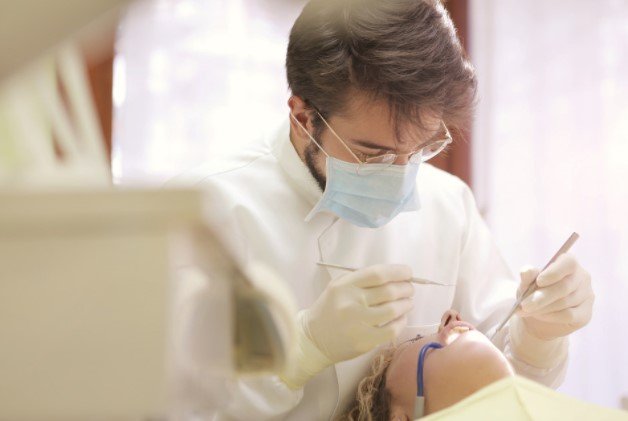 Pessoa, que aparenta ser um dentista, consultando a boca de um paciente deitado em maca hospitalar - Metropoles