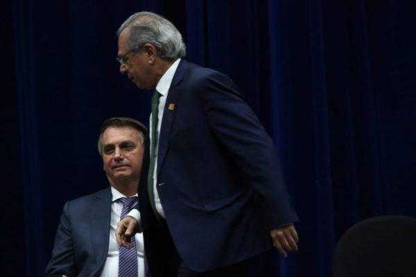 Bolsonaro assistindo a Guedes se levantar em cerimônio no Inmetro. Ambos usam terno - Metrópoles