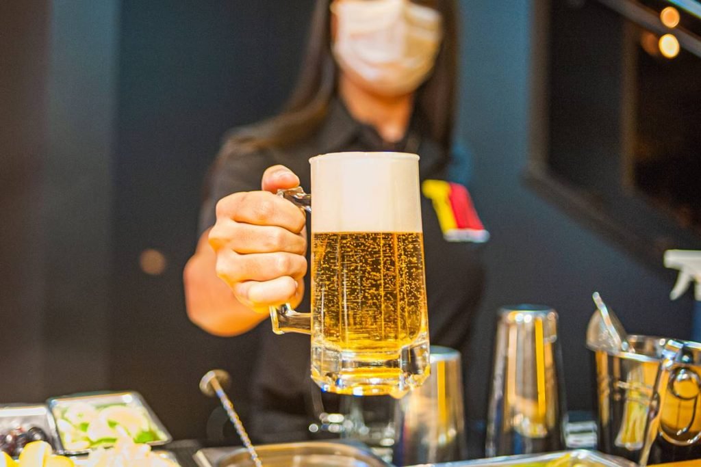 na foto temos um copo de chope segurado por uma mulher desfocada ao fundo