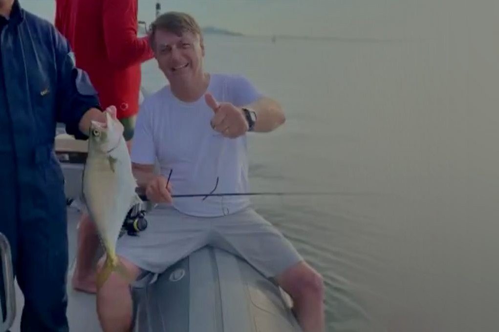 Bolsonaro passando as férias na praia. Ele usa camiseta clara e está segurando uma vara de pescar dentro de um barco enquanto alguém ao lado dele está com um peixe nas mãos- Metrópoles