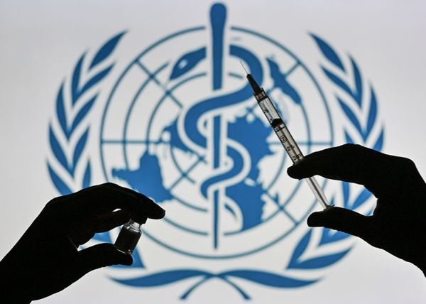 Bandeira da Organização Mundial da Saúde (OMS) atrás de duas mãos. Uma segura uma seringa e outra segura recipiente com liquido transparente