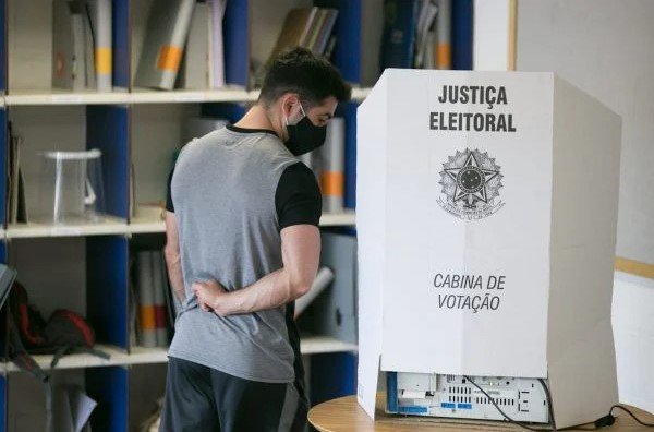 Homem vestido com blusa cinza e calça preta ao lado de uma urna eleitoral - metrópoles