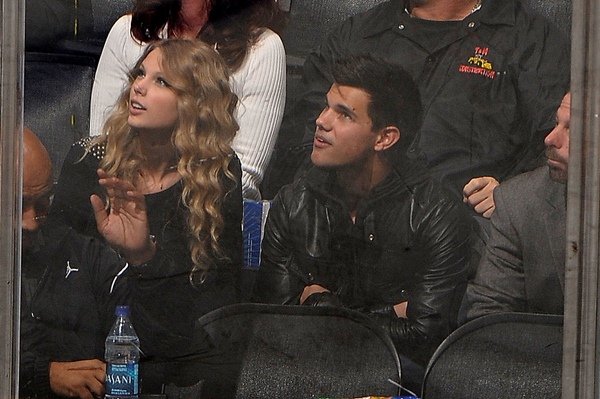 Taylor Swift e Taylor Lautner. Eles estão em um jogo esportivo e usam roupas de frio escuras - Metrópoles