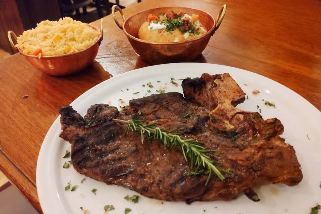 na foto temos um prato com um pedaço de carne e ao fundo duas cumbucas de cobre