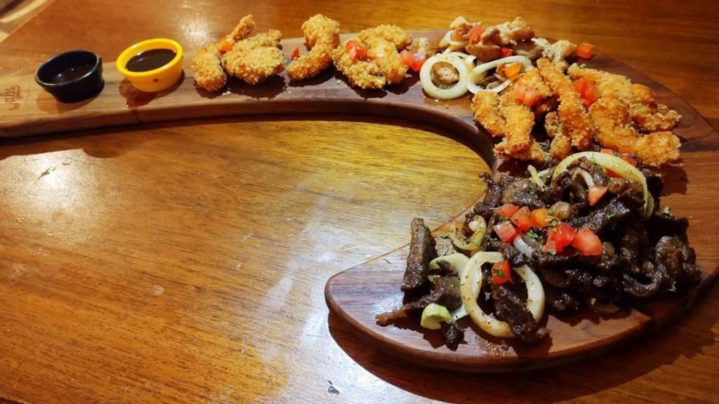 na foto vemos uma tabua de madeira em formato de jota com carnes em cima