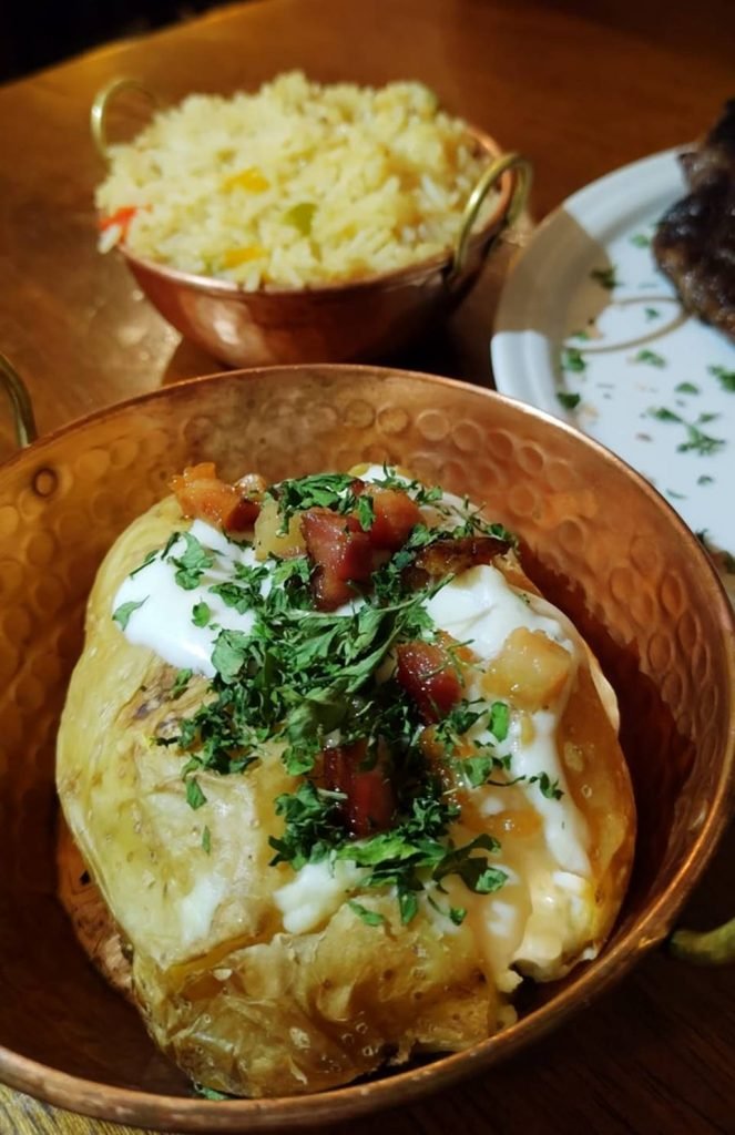 na foto temos uma cumbuca de cobre com uma bata recheada e coberta de bacon e ao fundo outra cumbuca com arroz