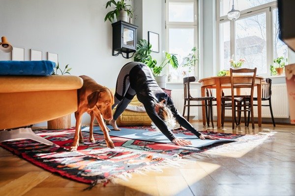 Mulher praticando ioga e há um cachorro ao lado dela. Ela tem cabelos loiros e usa roupa escura