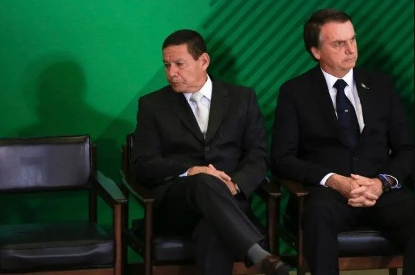 Bolsonaro e Mourão olham para lados opostos em cerimônia.  Eles vestem terno e gravata e tem cabelos escuros - Metrópoles