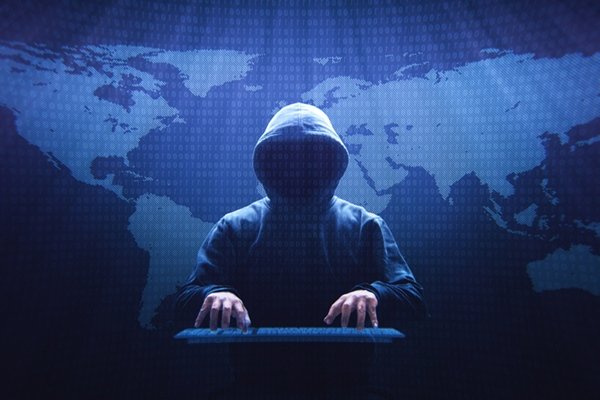 Pessoa com capuz de frente digita num teclado. ao Fundo, uma luz azul com mapa mundi - Metrópoles