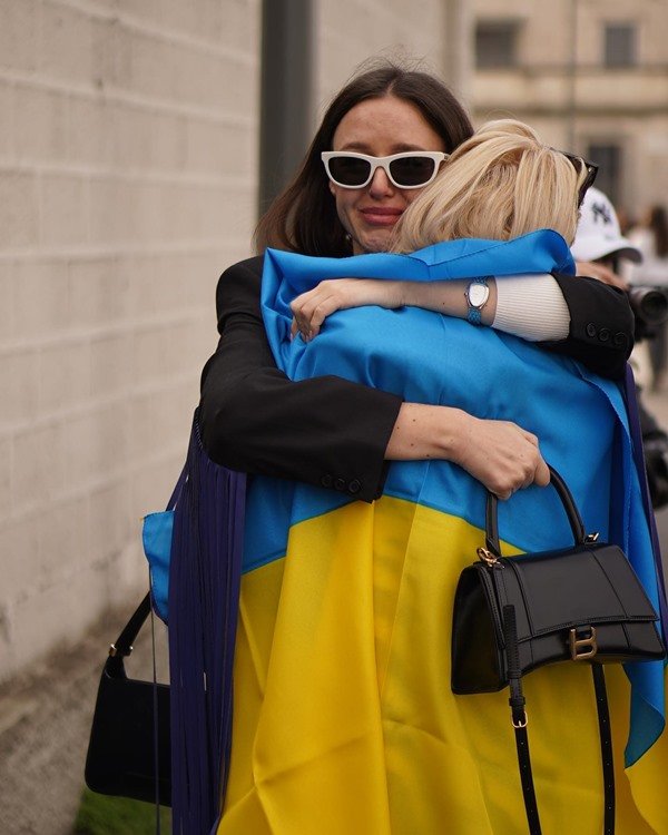 Duas mulheres se abraçando e chorando. Uma tem o cabelo branco e a outra tem o cabelo preto. Uma delas está com a bandeira da Ucrânia sobre os ombros.