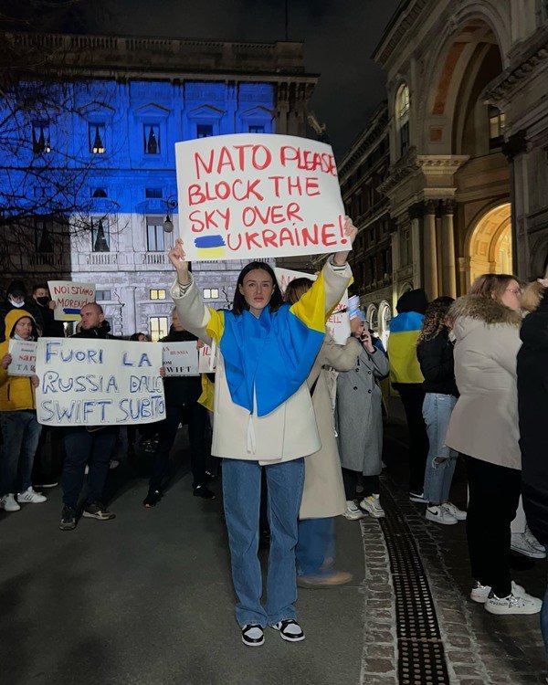 Mulher branca de cabelo preto segura bandeira da Ucrânia e cartaz pedindo o fim da invasão da Rússia no país. Ao fundo é possível ver mais pessoas com cartazes no que parece ser uma manifestação.