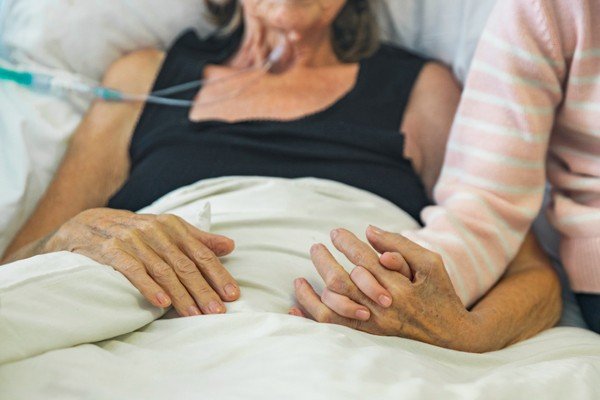 Woman lying in hospital bed holds the hand of a visitor - Metropolis