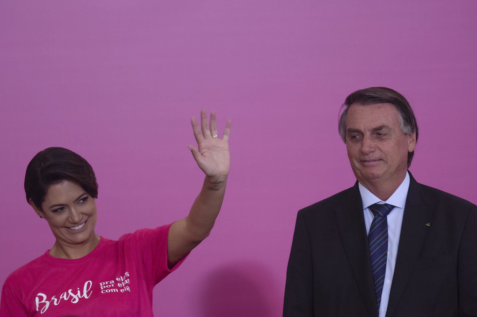 Michelle e Jair Bolsonaro em cerimônia de comemoração do Dia da Mulher no Palácio do Planalto. Ela acena para a plateia e usa rosa - Metrópoles