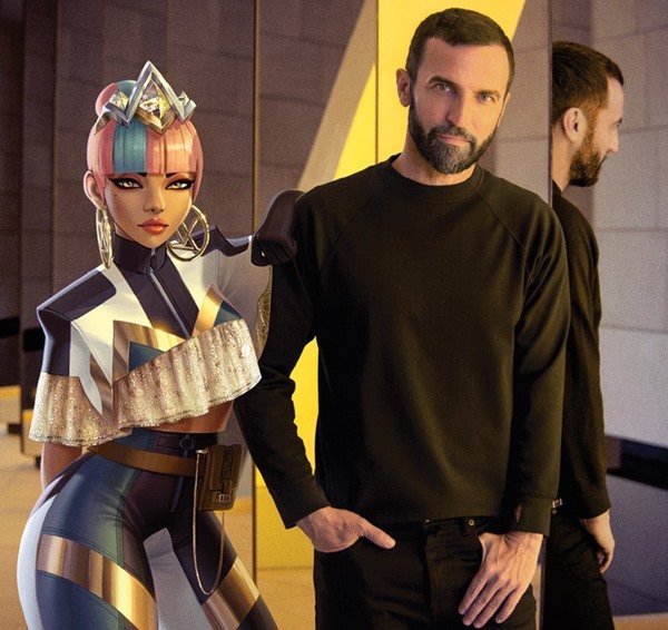 O estilista Nicolas Ghesquière posando ao lado de uma personagem virtual do videogame League of Legends. Ele veste preto e a personagem possui o cabelo rosa e usa roupa dourada e azul - Metrópoles