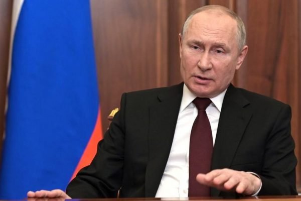 Presidente russo Vladimir Putin. Ele está sentado em frente à uma mesa com a bandeira russa atrás - Metrópoles