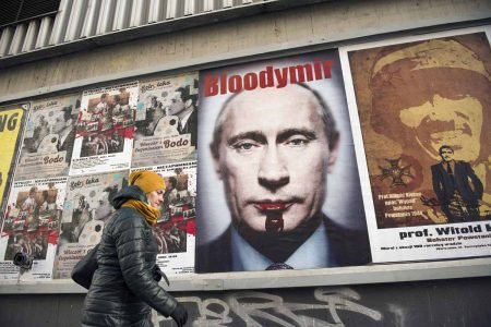 Mulher passa por cartaz de repúdio ao presidente russo Vladimir Putin escrito "Bloodymir" com sangue escorrendo da boca do líder em Varsóvia, Polônia - Metrópoles