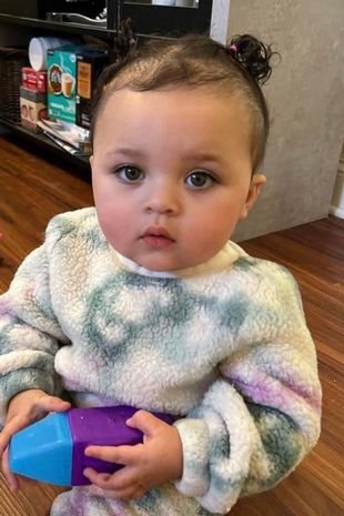 Bebê branca de cabelo escuro e olho azul, com roupa de frio e segurando um brinquedo