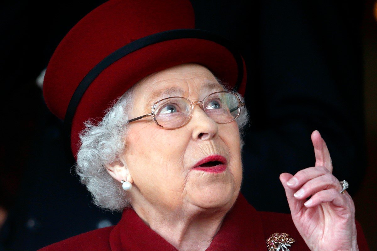Foto colorida. Rainha Elizabeth com chapéu e roupa vermelha. Ela está com o dedo levantado