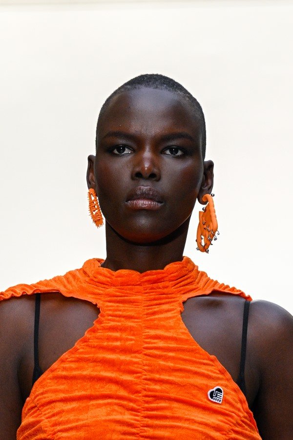 Mulher negra e careca usando brinco e vestido laranja. As peças são da marca Ester Manas.
