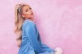A socialite Paris Hilton de perfil, com sua cachorrinha, faz pose e olha para câmera com agasalho azul de sua própria linha, em fundo rosa - Metrópoles