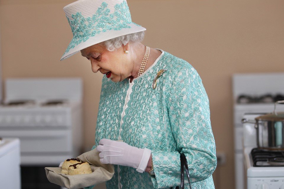Foto colorida. Rainha Elizabeth com vestido e chapéu nas cores verde menta e branco. Ela segura um pão