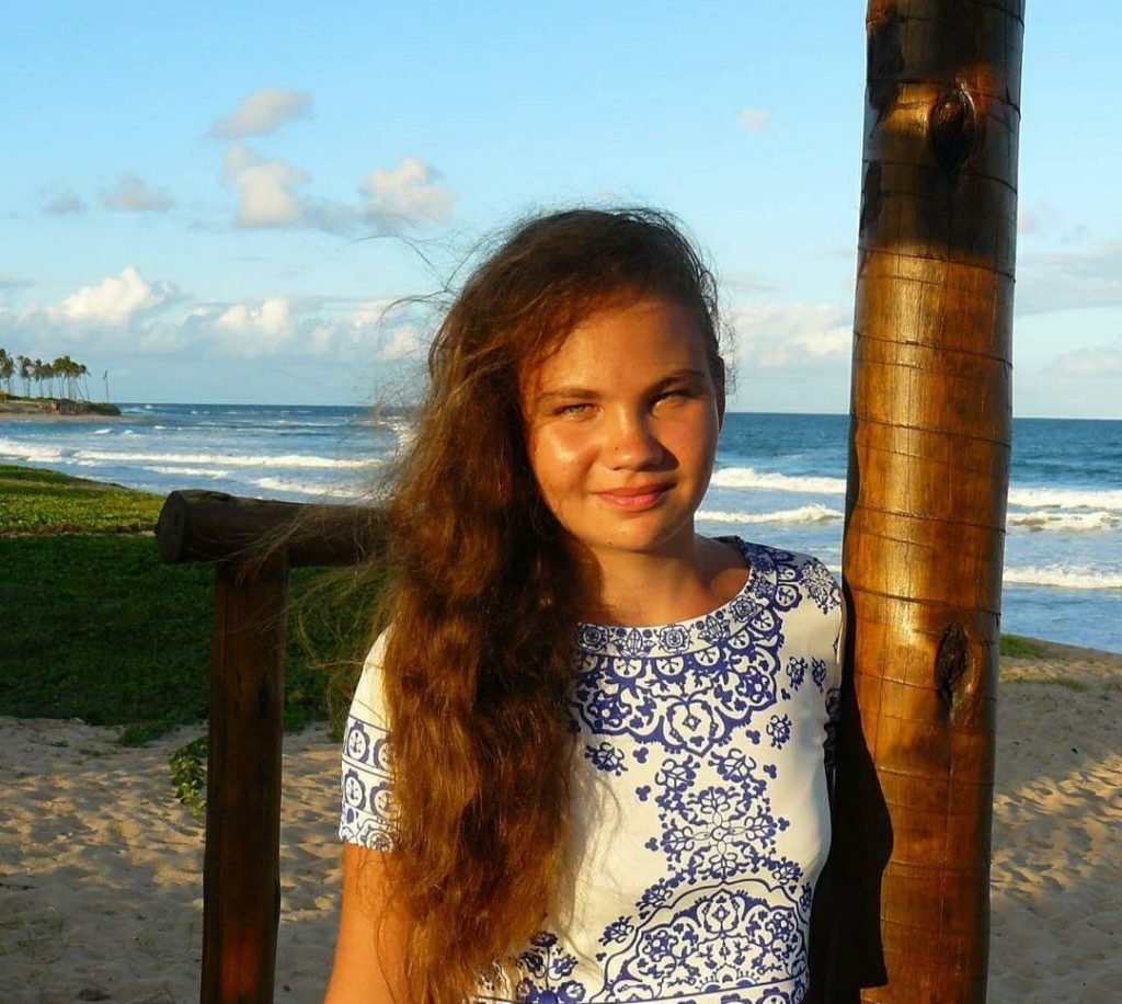 Menina com cabelo cacheado encostada em tronco de madeira e praia ao fundo